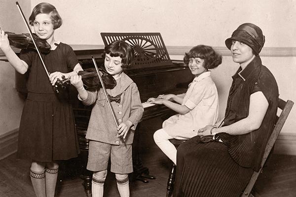 woman wearing hat with child at piano and two children with violins